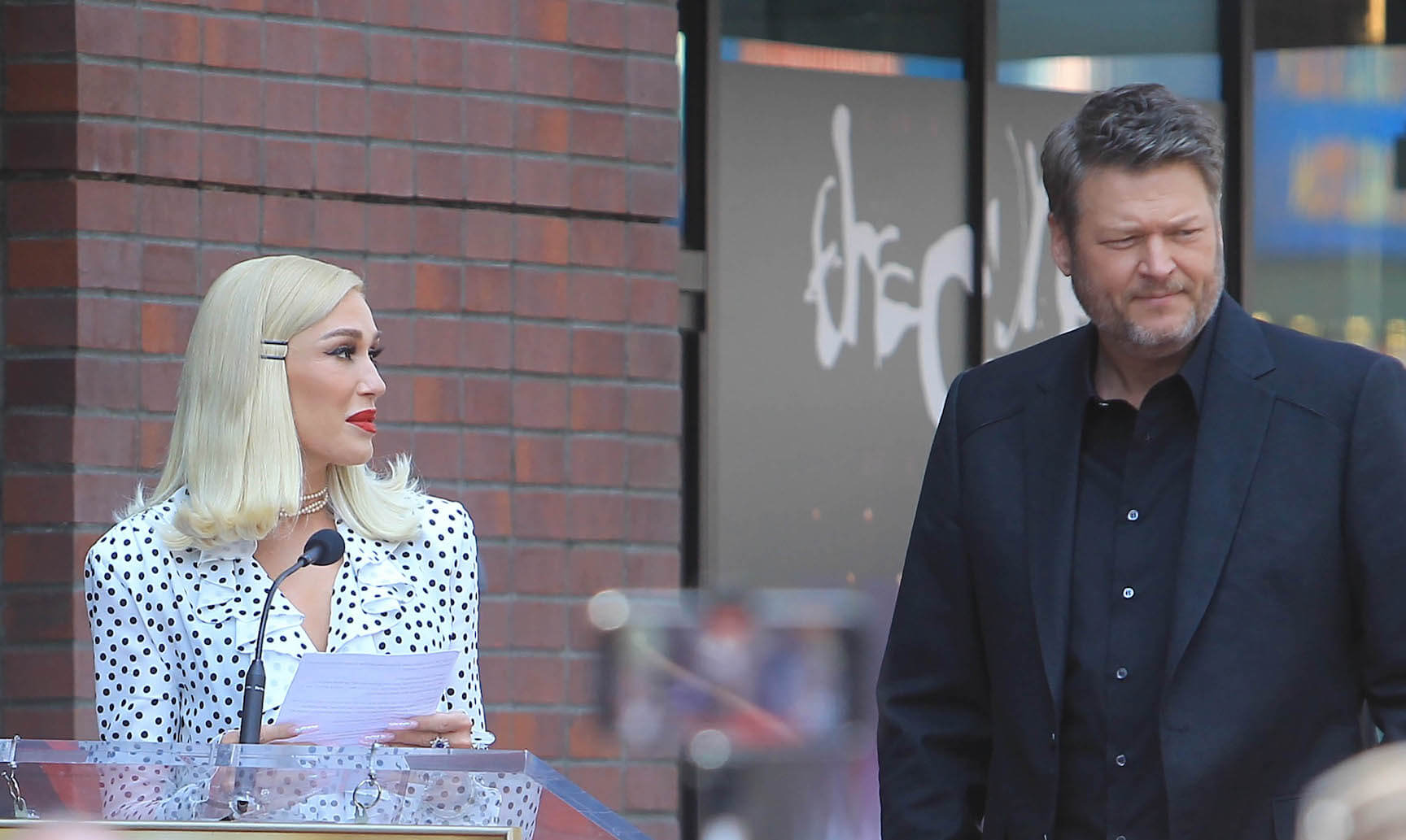 Gwen Stefani and Blake Shelton standing next to each other while Stefani gives a speech in Los Angeles
