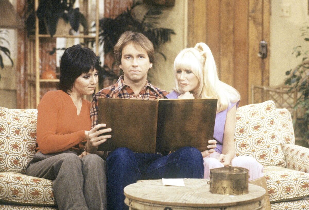 The cast of 'Three's Company' John Ritter, Suzanne Somers, and Joyce Dewitt, sitting on a couch.