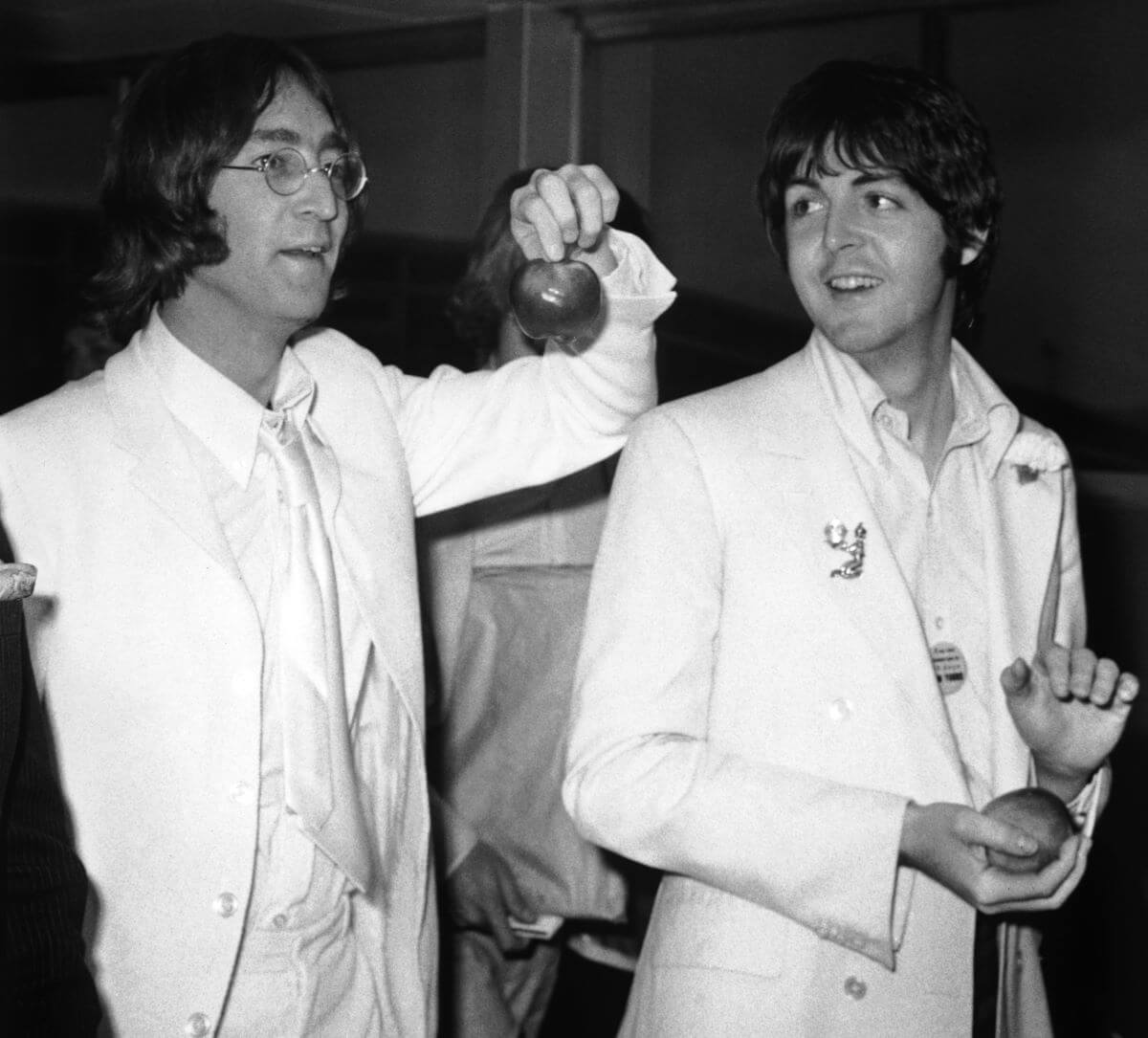 A black and white picture of John Lennon and Paul McCartney wearing white suits and holding apples.