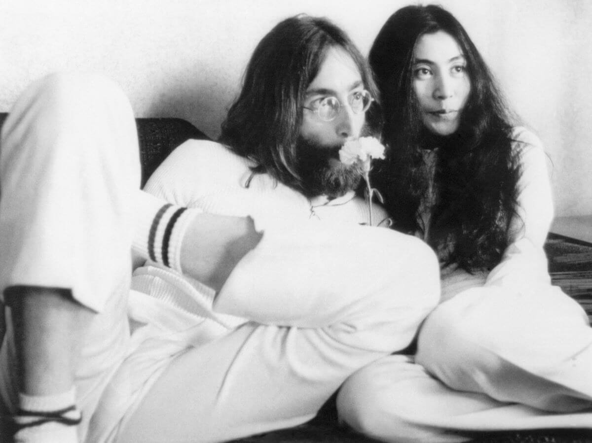 A black and white picture of John Lennon and Yoko Ono sitting on a mattress together. Lennon holds a flower up to his nose.