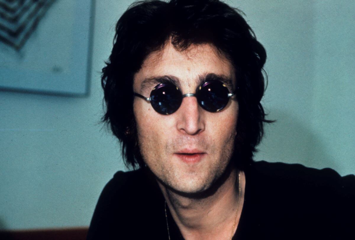 John Lennon wears a pair of dark circular sunglasses.