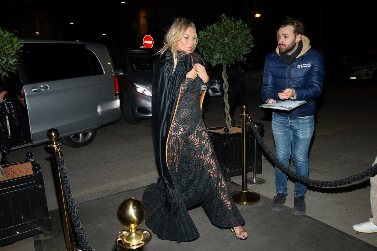 Kate Moss walking while wearing a black dress and coat at the Ritz.