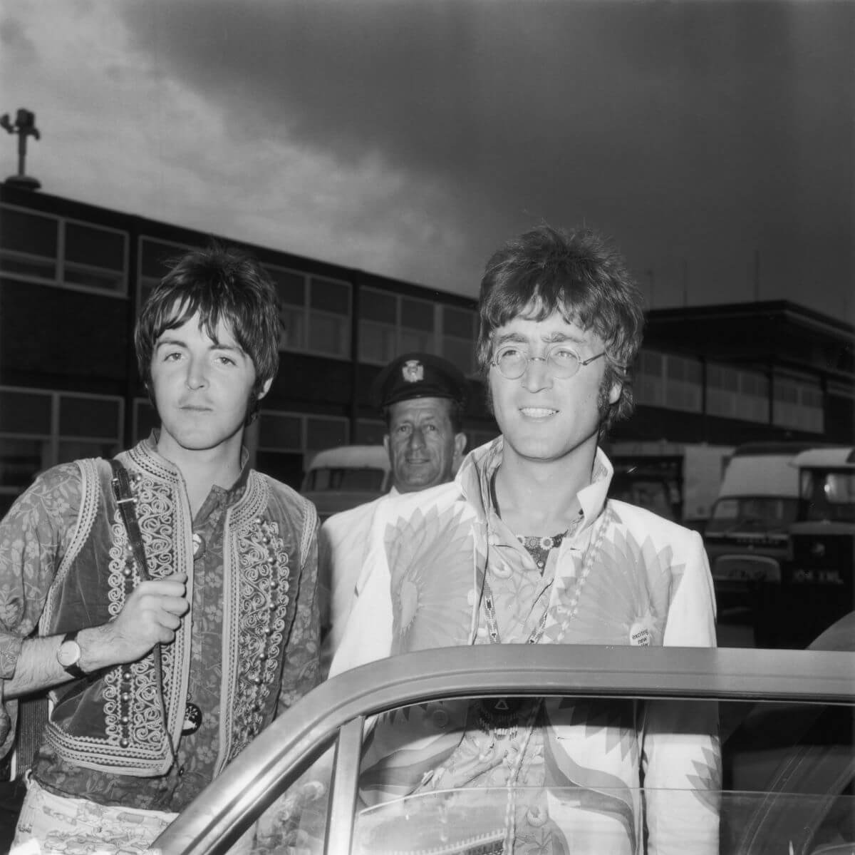 A black and white picture of John Lennon and Paul McCartney holding bags and standing behind an open car door.