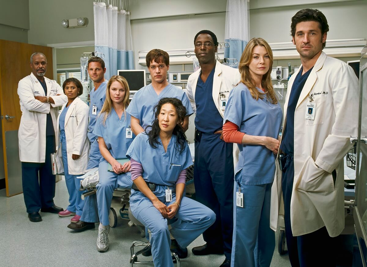 Patrick Dempsey posing alongside 'Grey's Anatomy' cast while wearing a white lab coat that covers a blue outfit.