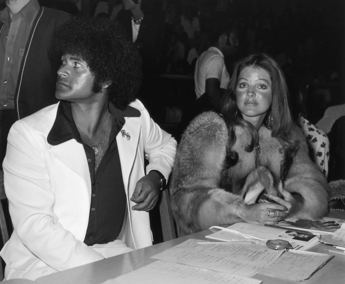 A black and white picture of Mike Stone sitting at a table with Priscilla Presley, who wears a fur coat.