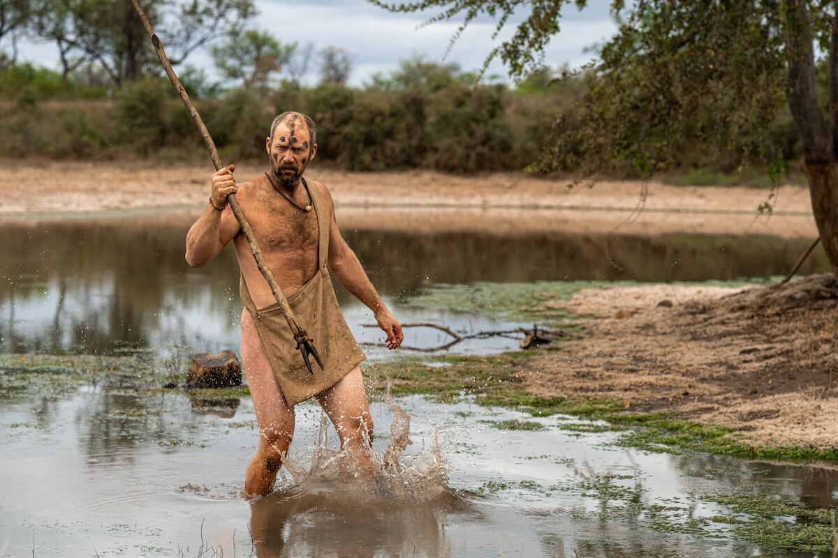 Cole from 'Naked and Afraid' walking in the pond, holding a large walking stick.