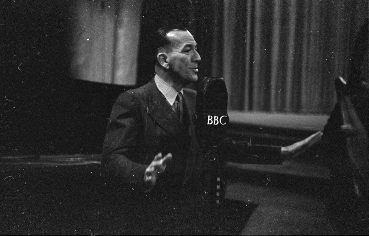 A black and white picture of Noel Coward speaking into a microphone.