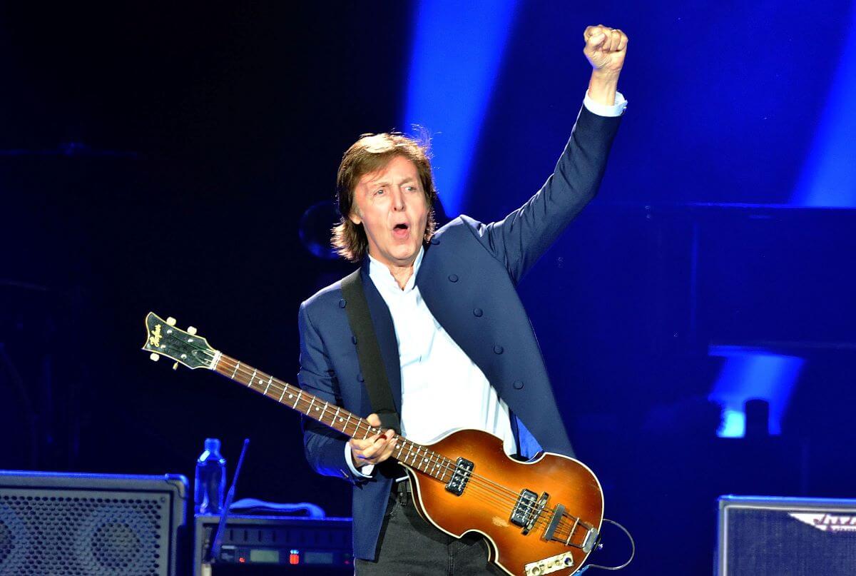 Paul McCartney holds a guitar and lifts his fist into the air.