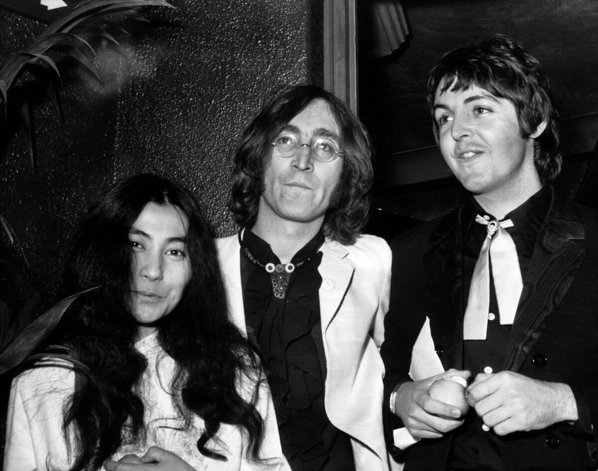 A black and white picture of Yoko Ono, John Lennon, and Paul McCartney. McCartney holds an apple.