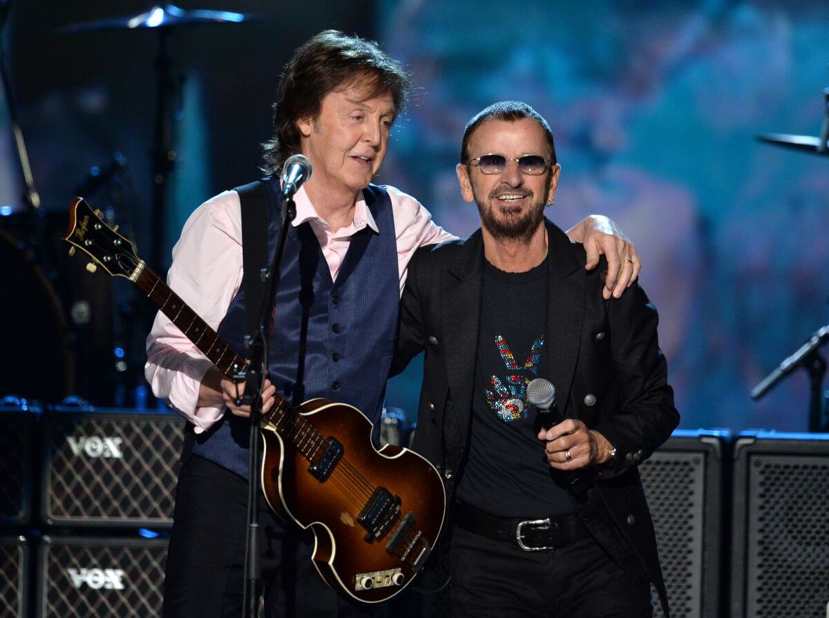Paul McCartney holds a guitar and stands with his arm around Ringo Starr's shoulders.