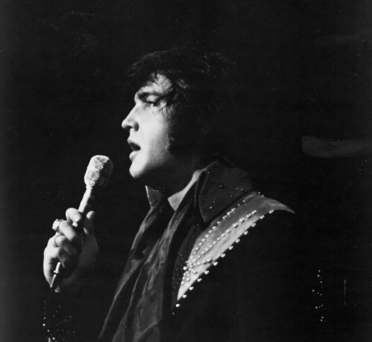 A black and white picture of Elvis Presley singing into a microphone while in side profile.
