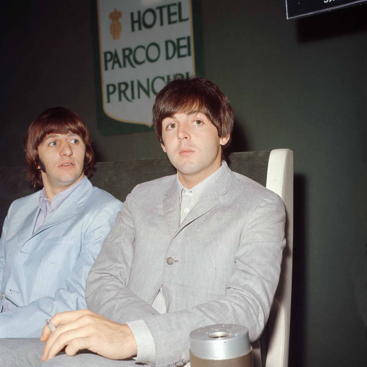 Ringo Starr and Paul McCartney sit next to each other on the same side of a table. Starr wears a blue suit and McCartney wears a gray one.