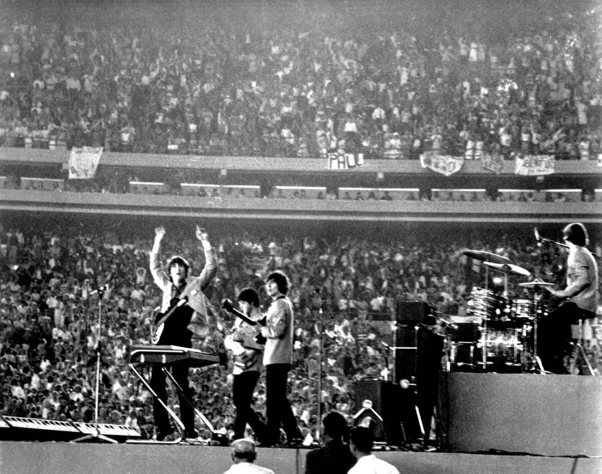 A black and white picture of John Lennon, Paul McCartney, George Harrison, and Ringo Starr onstage at Shea Stadium. Lennon lifts his arms in the air and Starr sits at a drum set.