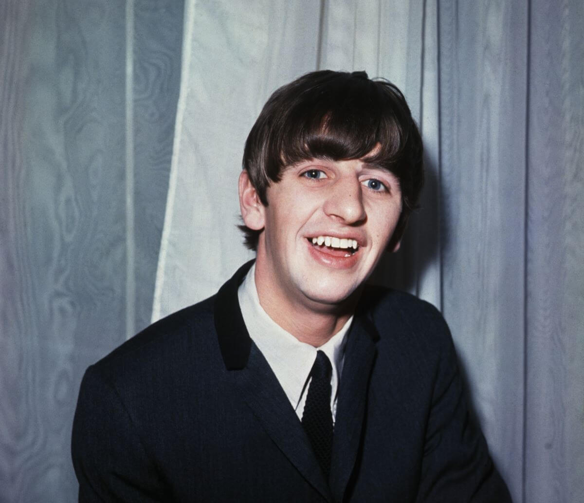 Ringo Starr wears a suit and sits in front of a gray curtain.