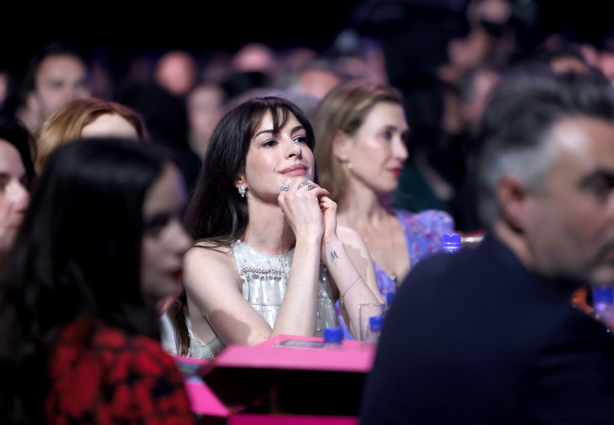 Anne Hathaway sits with her hands under her chin in a crowd of people.