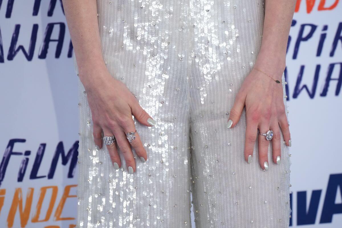 Anne Hathaway's hands with painted silver nail polish and silver rings. She holds them against her silver jumpsuit.