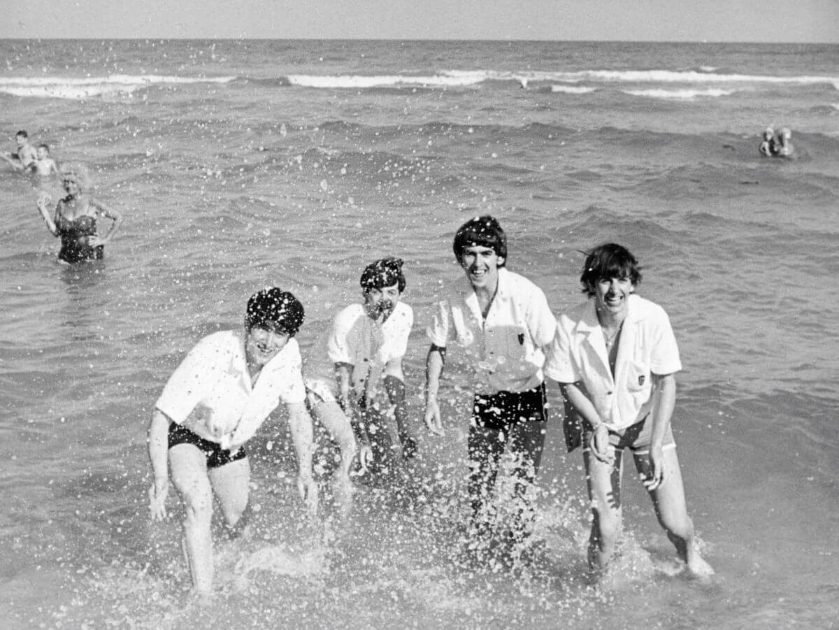 A black and white picture of The Beatles standing in the ocean. They all wear white shirts.