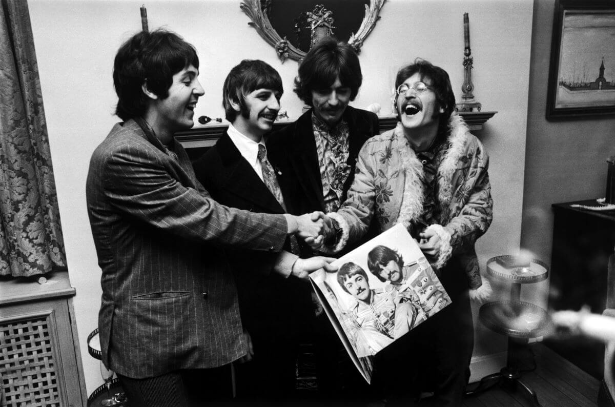 A black and white picture of Paul McCartney and John Lennon laughing while shaking hands. Ringo Starr and George Harrison look on. 