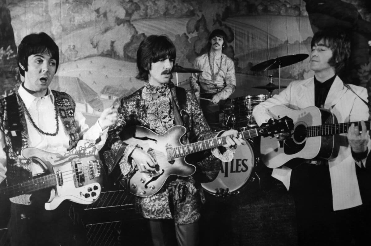 A black and white picture of Paul McCartney, George Harrison, and John Lennon holding guitars. Ringo Starr sits at a drum set behind them.