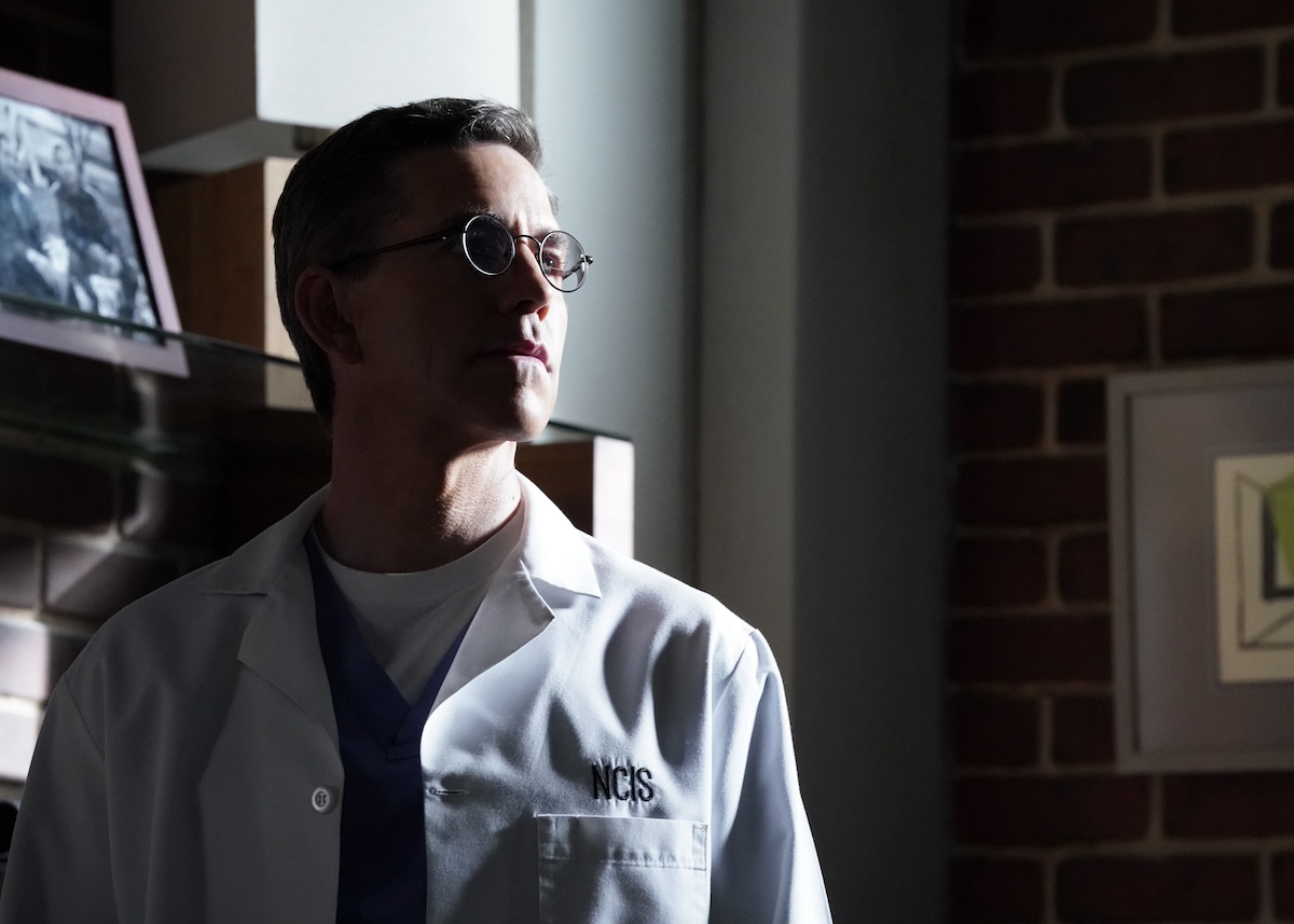 Brian Dietzen in a lab coat and standing partly in shadow in 'NCIS'