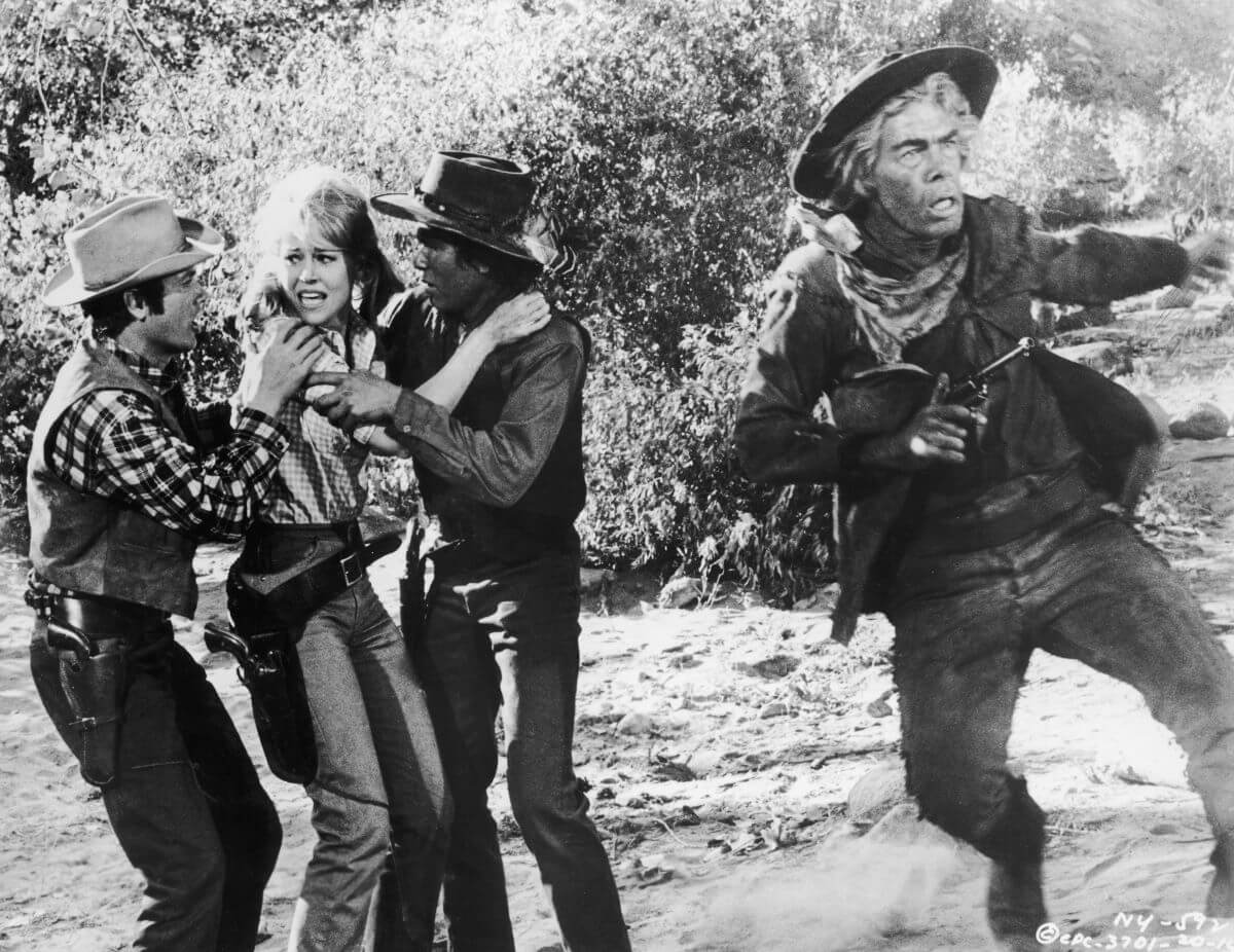 A black and white still of Lee Marvin holding a gun and jumping in front of Jane Fonda and two other actors in 'Cat Ballou.'