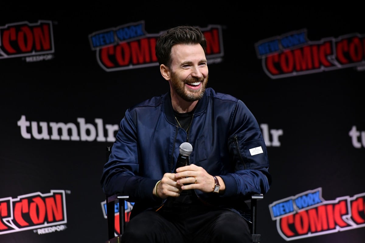 Chris Evans speaking at a New York comic con panel wearing a coat while smiling.