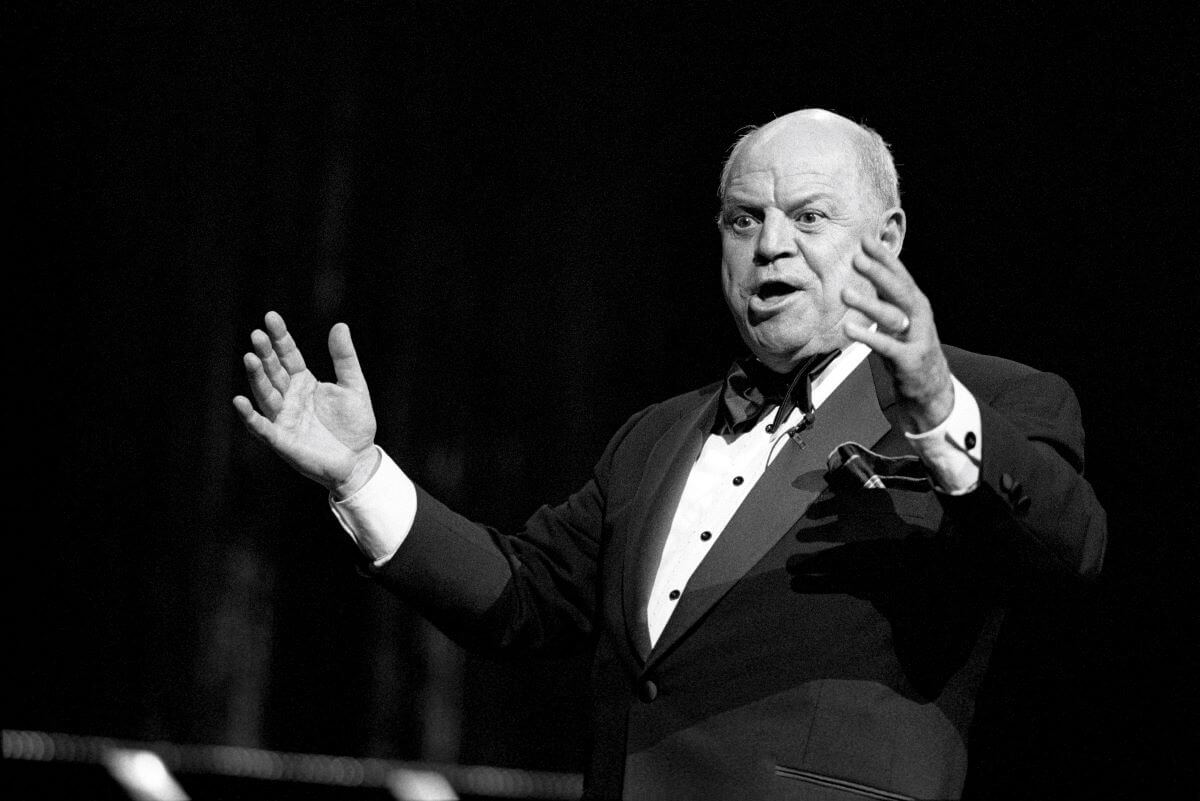 A black and white picture of Don Rickles wearing a tuxedo and lifting his arms while he speaks.