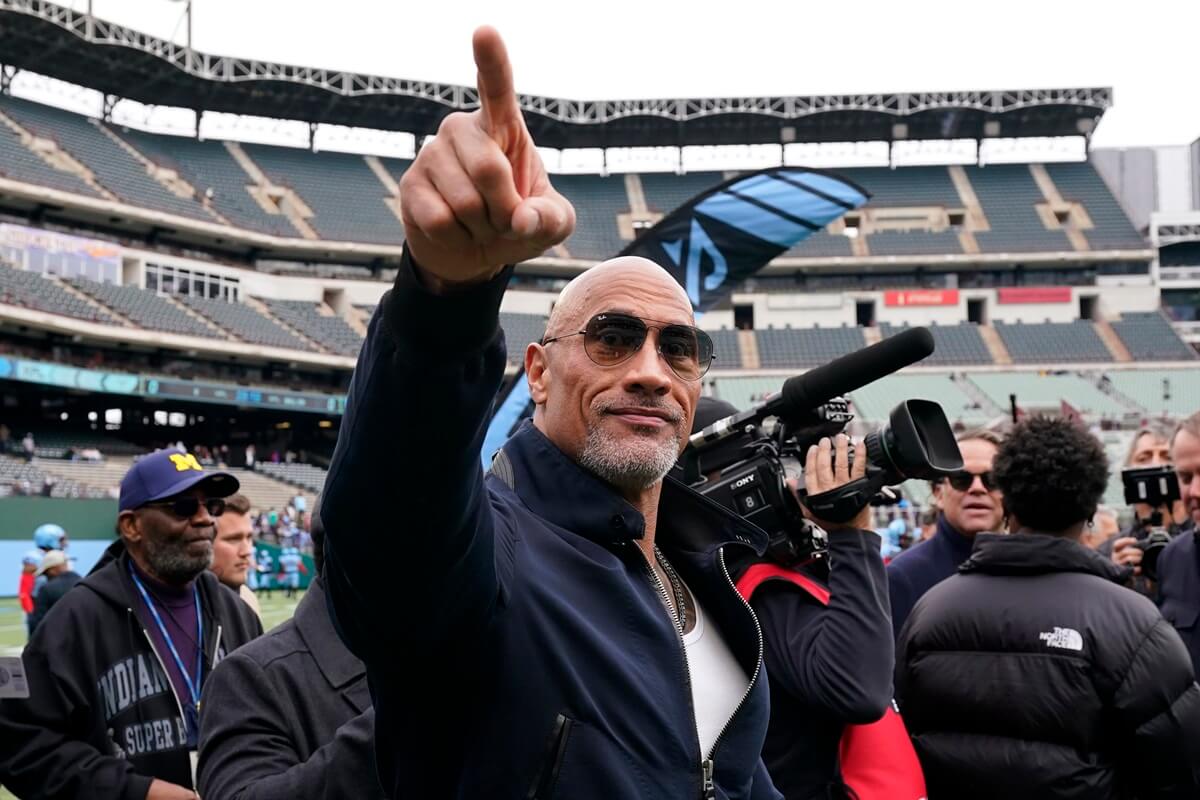 Dwayne Johnson posing at a football stadium wearing a black sweater and shades.