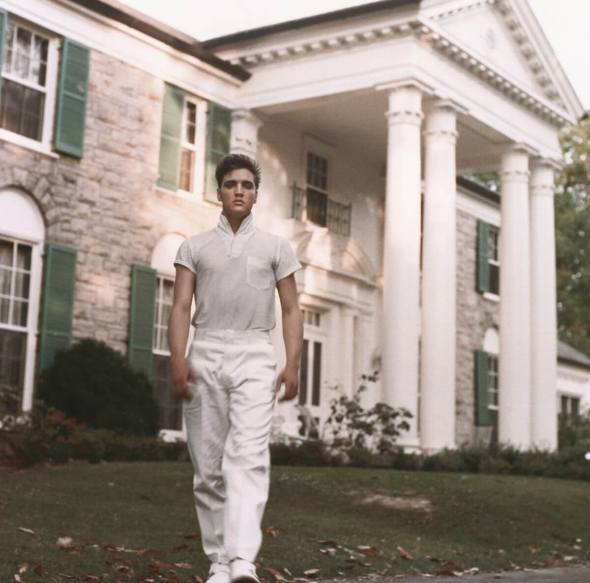 Elvis Presley wears a white shirt and pants and walks in front of Graceland.