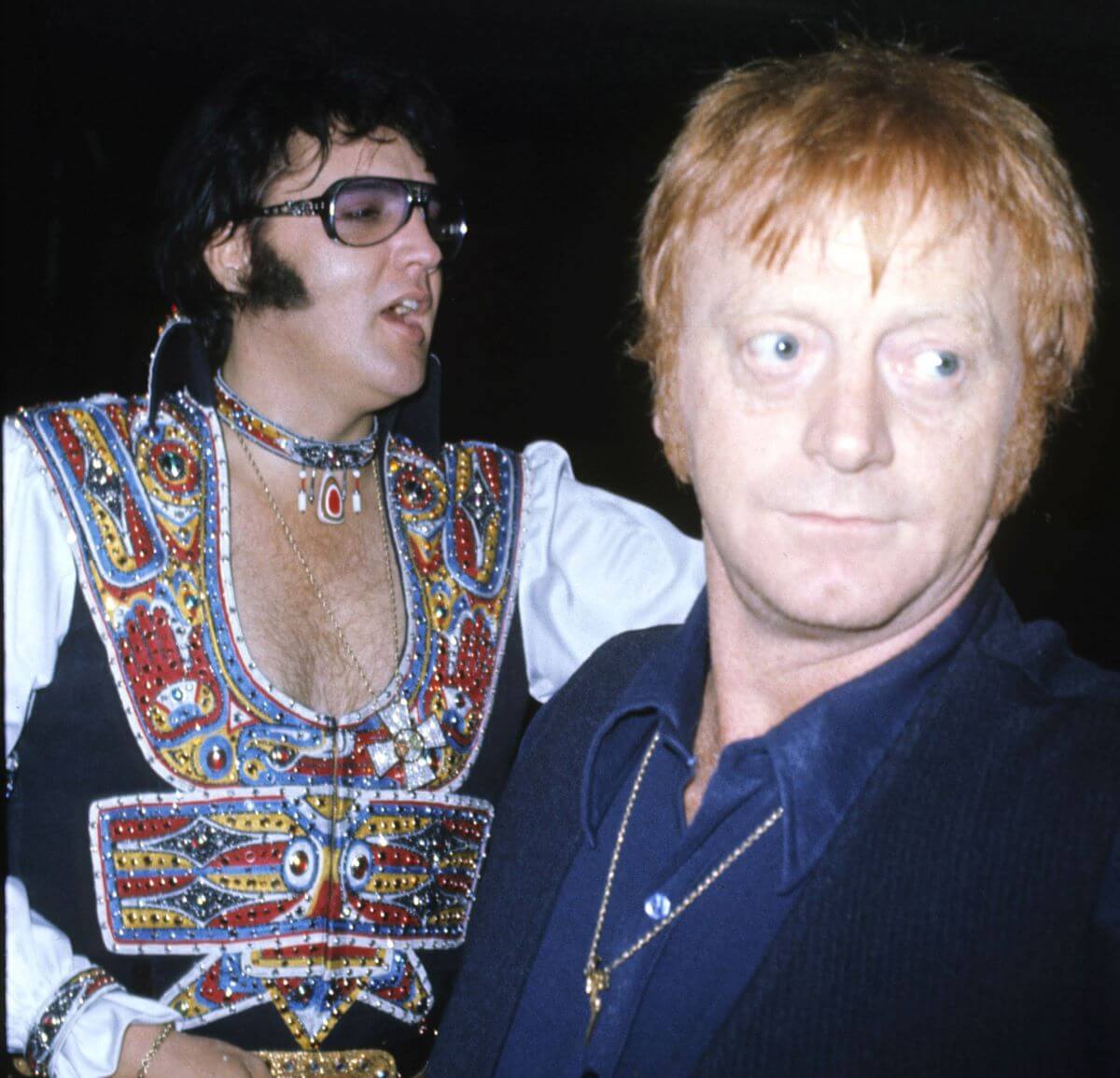 Elvis wears a bejeweled jumpsuit and sunglasses and stands behind his bodyguard, Red West.