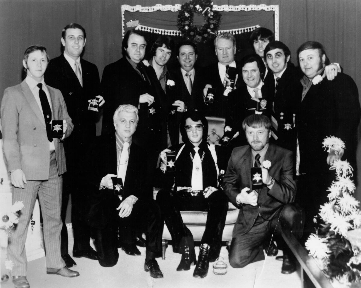 A black and white picture of Elvis sitting in a chair surrounded by his entourage. (L-R, standing) Billy Smith, former sheriff Bill Morris, Lamar Fike, Jerry Schilling, Sheriff Roy Nixon, Vernon Presley, Charlie Hodge, Sonny West, George Klein, Marty Lacker. (L-R, front) Dr. George Nichopoulos, Red West. 