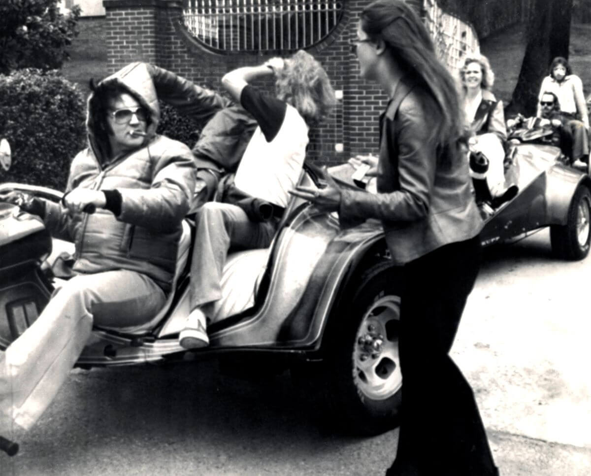 A black and white picture of Elvis riding a go kart out of the Graceland gates. A woman stands next to him.