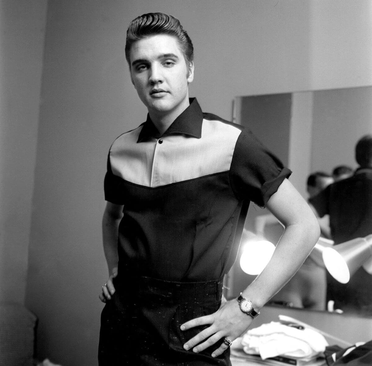 A black and white picture of Elvis wearing a polo shirt and standing with his hands on his hips.