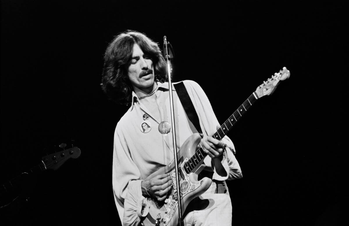 A black and white picture of George Harrison playing guitar while standing in front of a microphone.