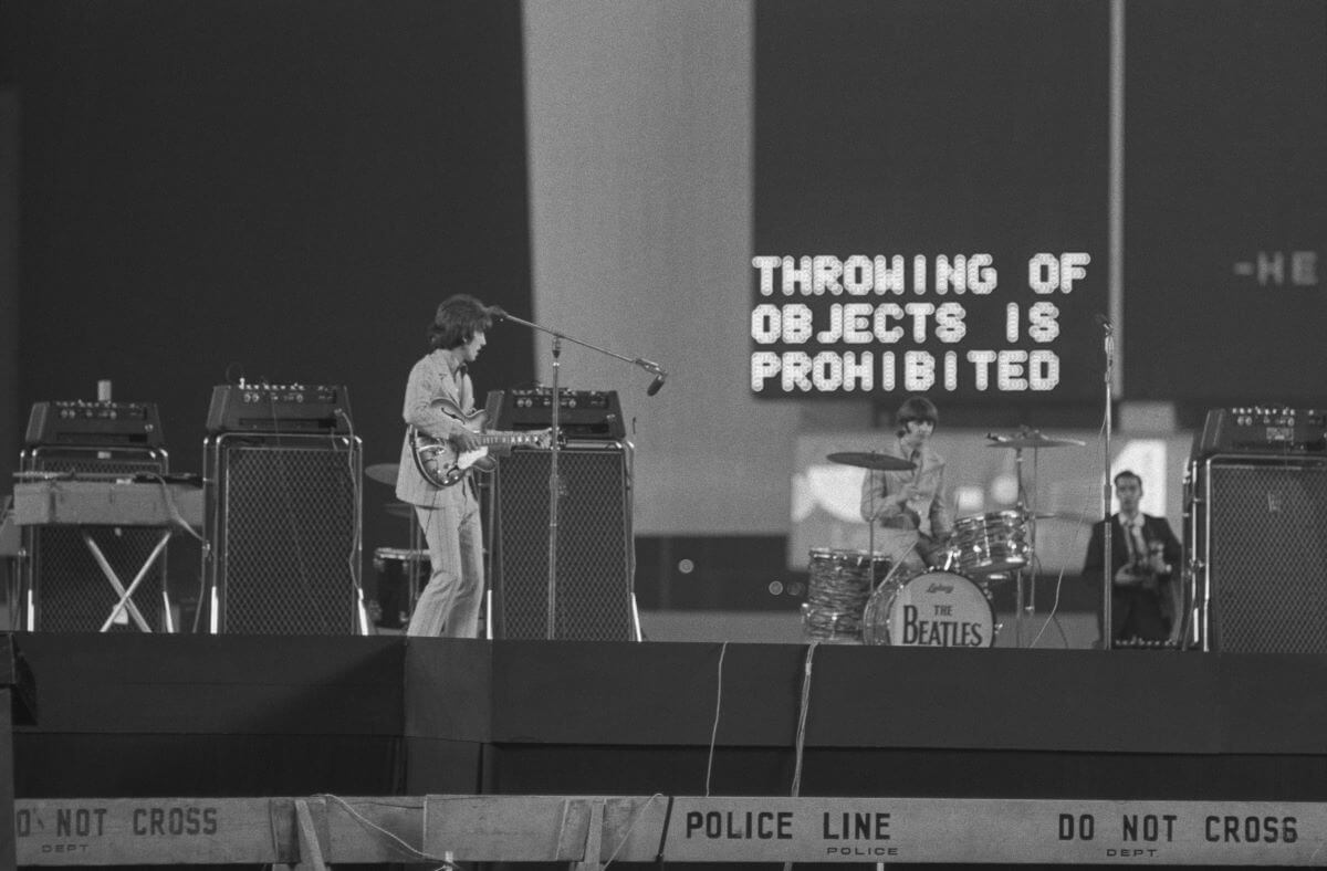 A black and white picture of George Harrison and Ringo Starr on stage in front of a sign that says, "Throwing of Objects Is Prohibited."