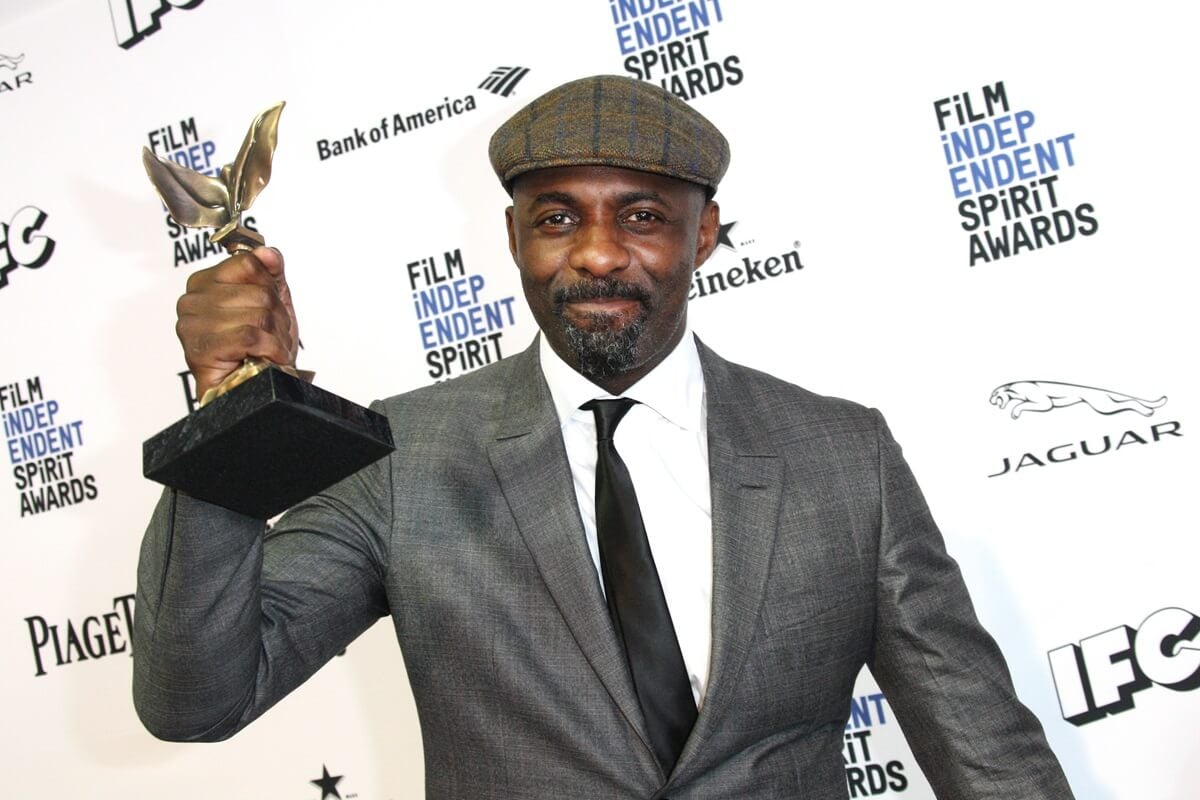 Idris Elba posing in a grey suit while holding an Independent Spirit Award.