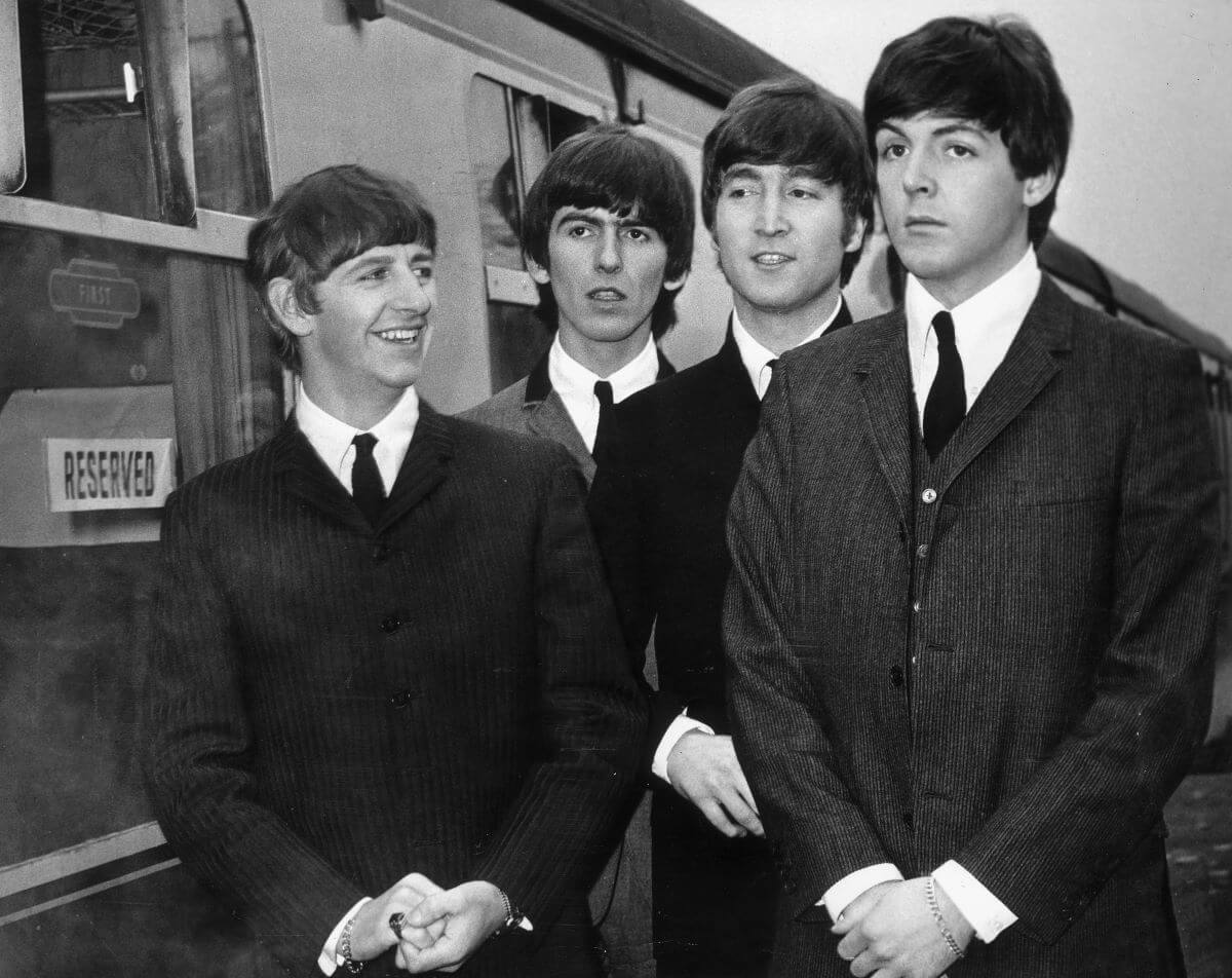 A black and white picture of Ringo Starr, George Harrison, John Lennon, and Paul McCartney of The Beatles standing next to a train. They wear suits.