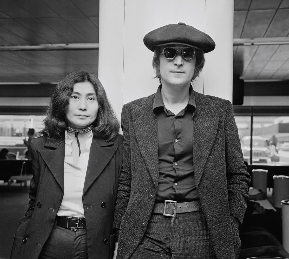 A black and white picture of Yoko Ono and John Lennon standing next to each other. Lennon wears a hat and sunglasses.