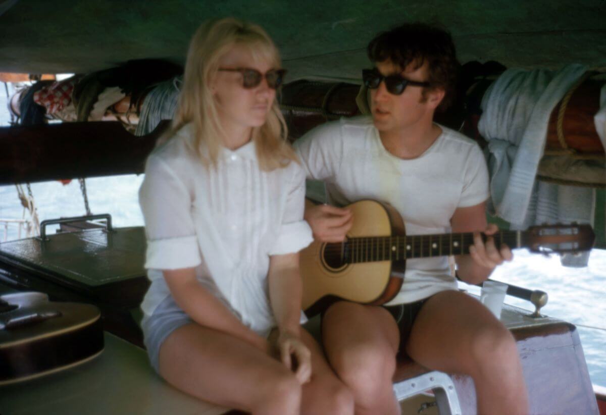 Cynthia Lennon sits next to John Lennon on a boat. They both wear white shirts and sunglasses, and he strums an acoustic guitar.
