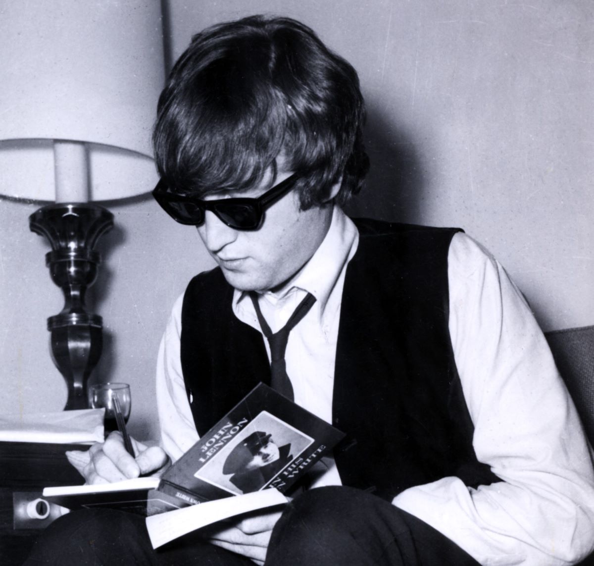 John Lennon wears sunglasses and a tie and signs copies of his book "In His Own Write."