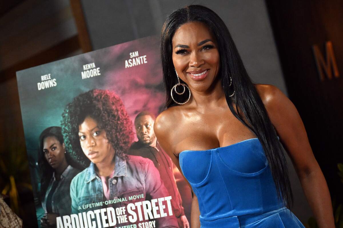 Wearing a blue velvet jumpsuit, Kenya Moore stands next to the promo image of her new Lifetime movie "Abducted Off The Street: The Carlesha Gaither Story"