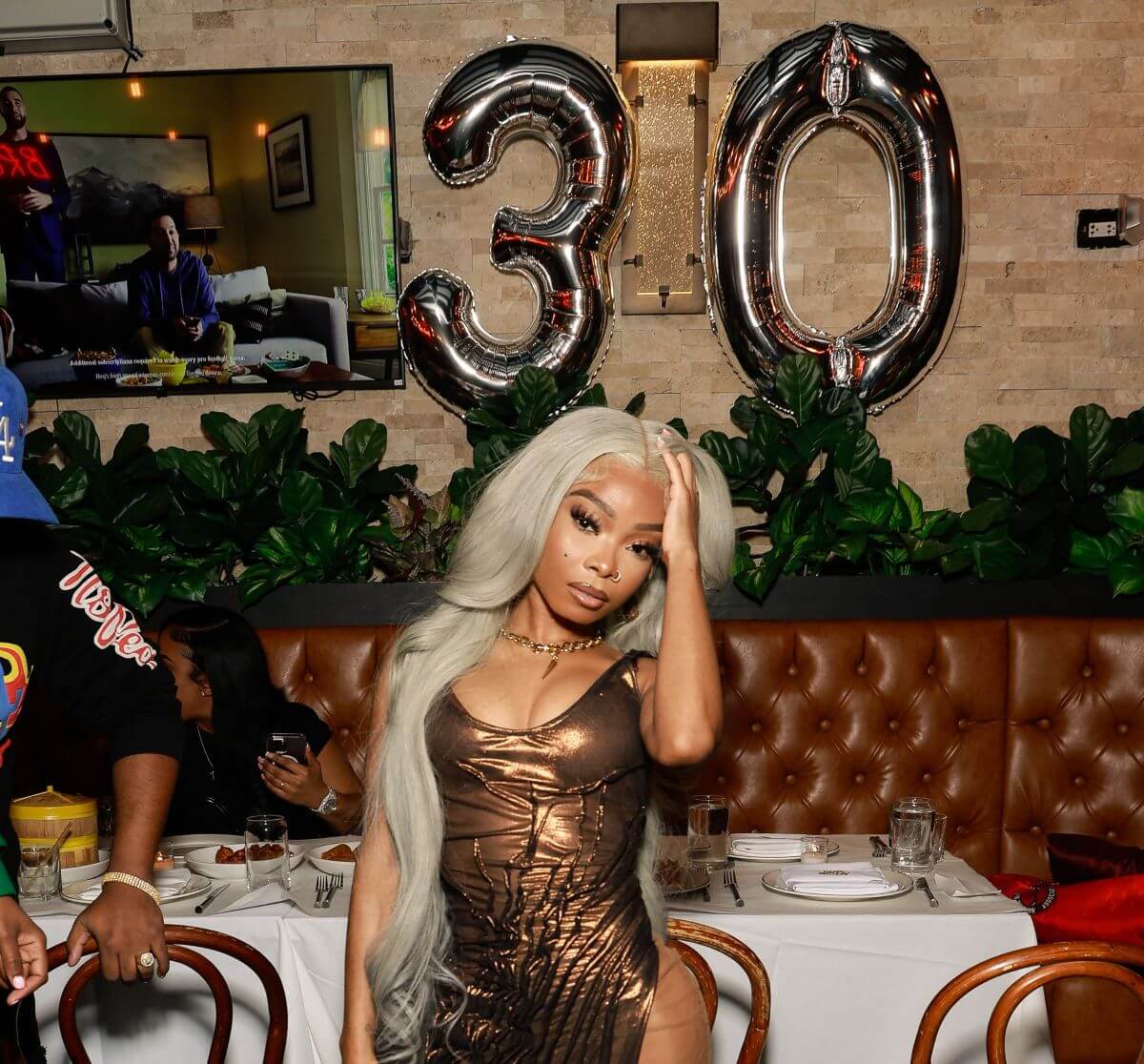 Lola Brooke runs her hand through her hair while standing in front of birthday balloons.