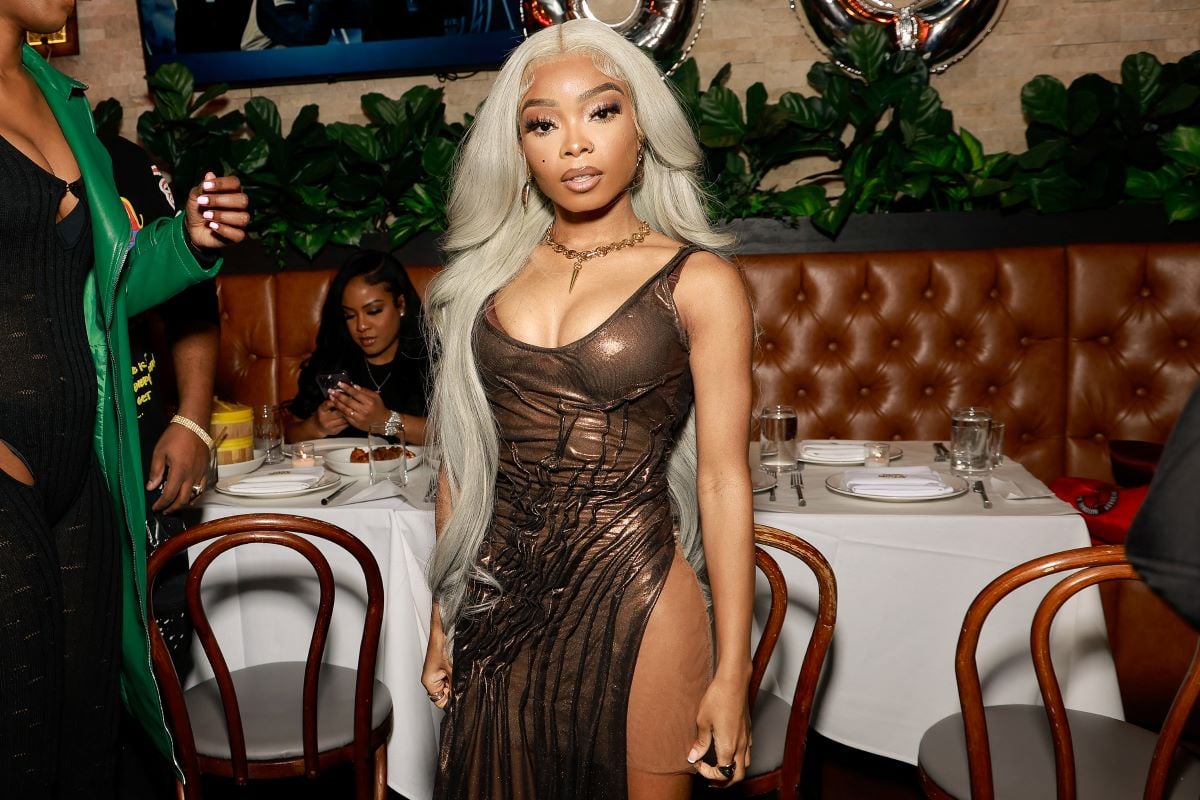 Lola Brooke stands in front of tables at her birthday celebration. She wears a brown dress.