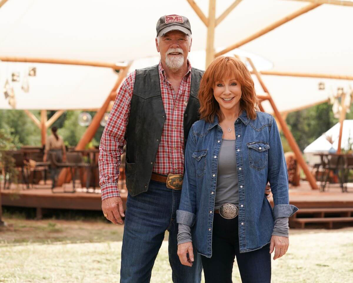 Reba McEntire wears a denim shirt and stands with her boyfriend, Rex Linn. He wears a plaid shirt, a vest, and a baseball hat.