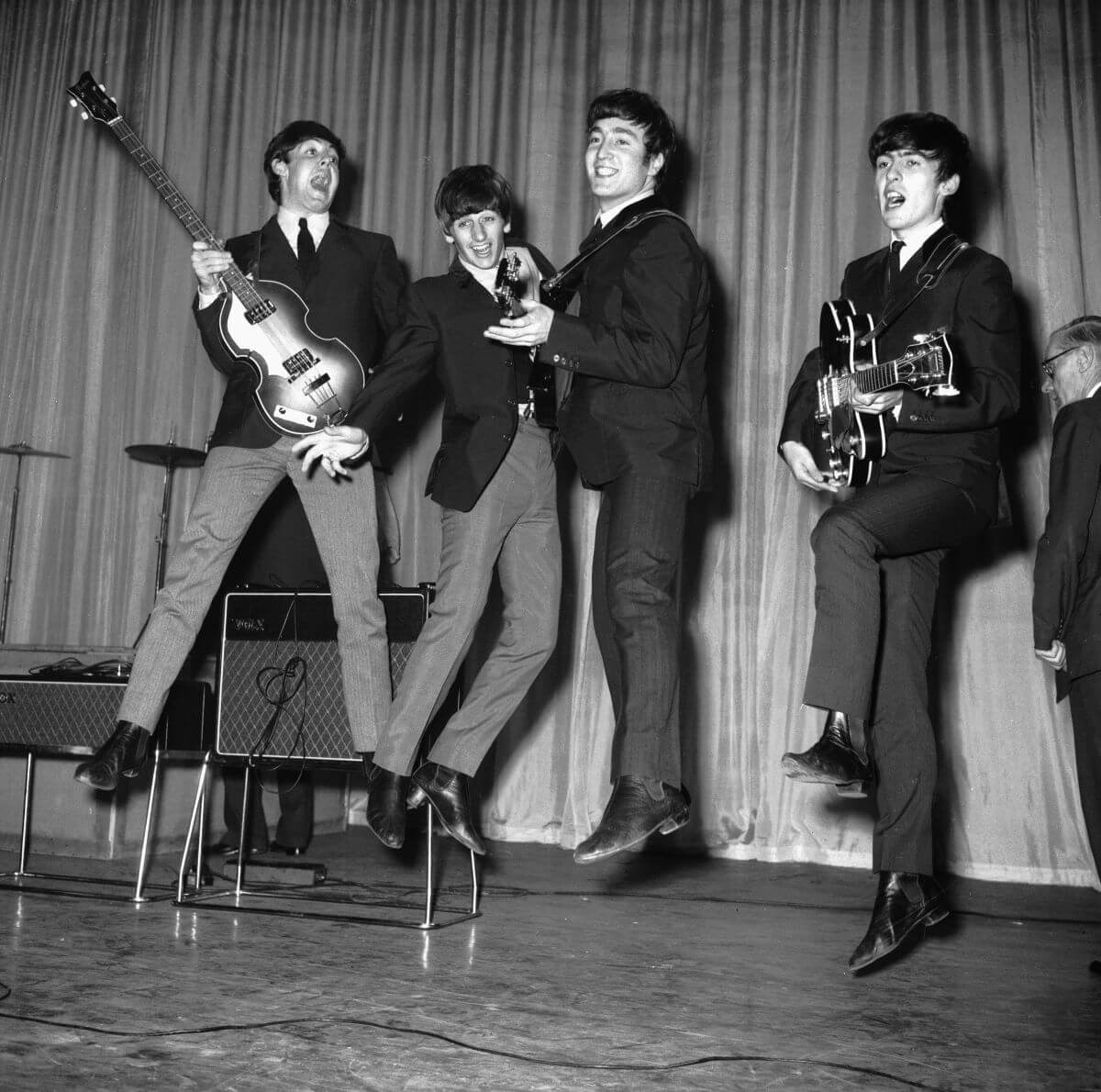 A black and white picture of The Beatles jumping up in the air while on a stage. Paul McCartney, John Lennon, and George Harrison hold guitars.