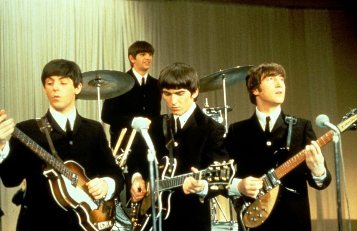 The Beatles play on a stage together. Paul McCartney, George Harrison, and John Lennon play guitars in a line and Ringo Starr plays the drums on a platform behind them.