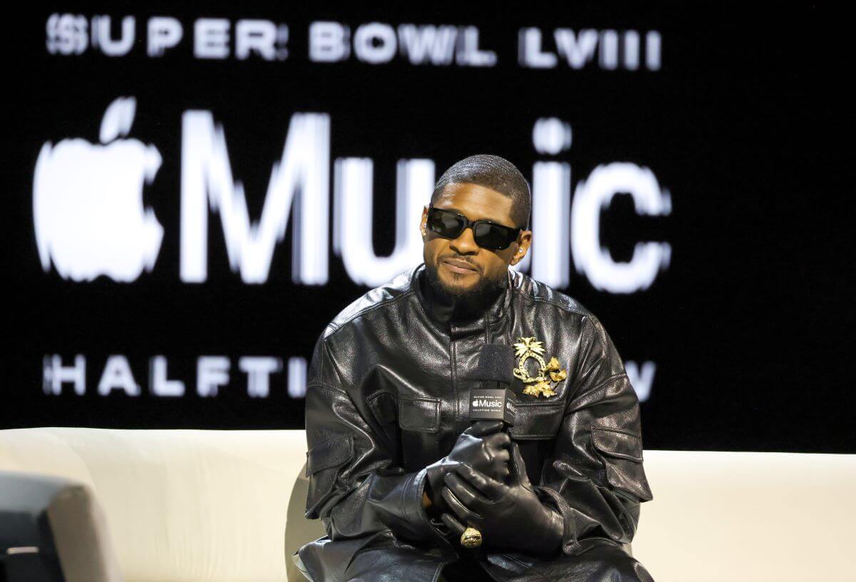 Usher wears sunglasses and a leather jacket. He sits in front of a sign for the Apple Music Super Bowl LVIII Halftime Show.