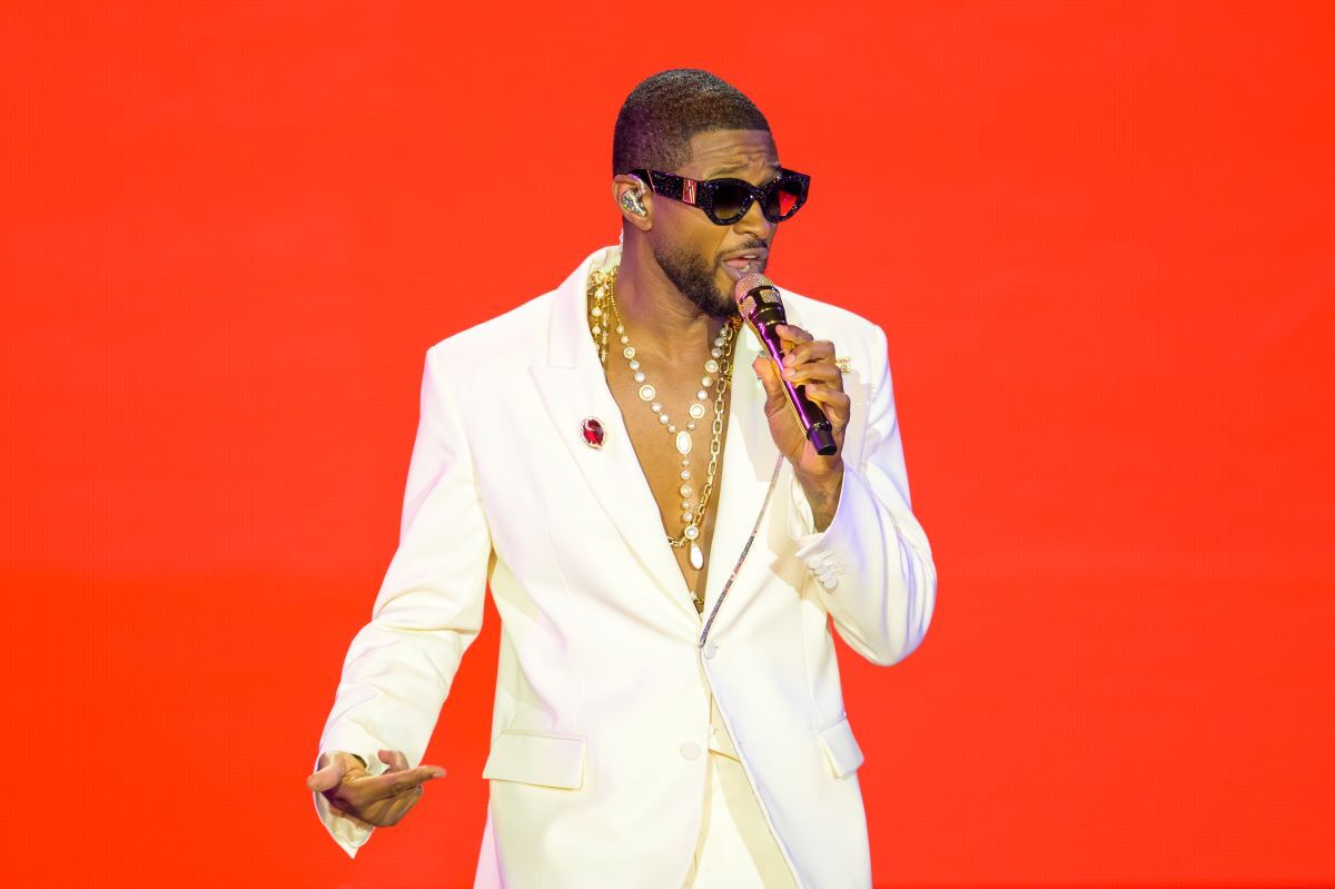 Usher wears a white suit, sunglasses, and a gold necklace. He sings into a microphone.