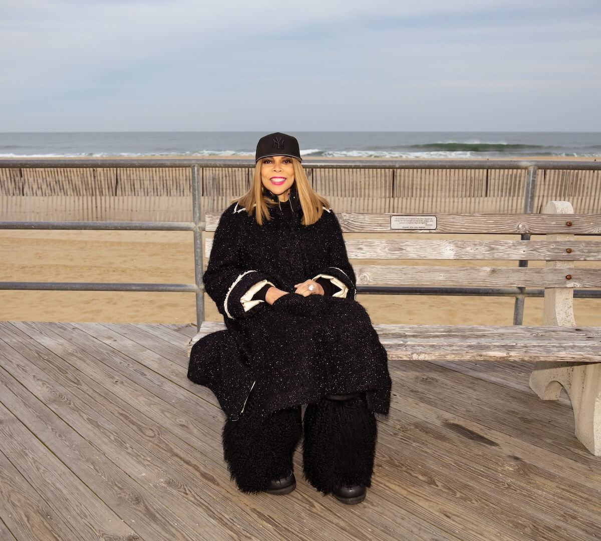 Wendy Williams, dressed in black, sitting on a bench in front of the ocean