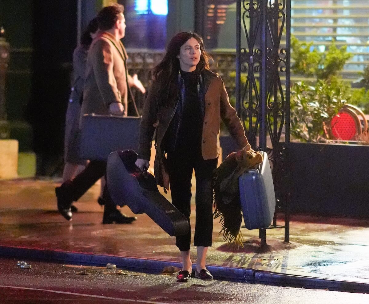 Monica Barbaro as Joan Baez, holding a guitar case and a suitcase, in NYC